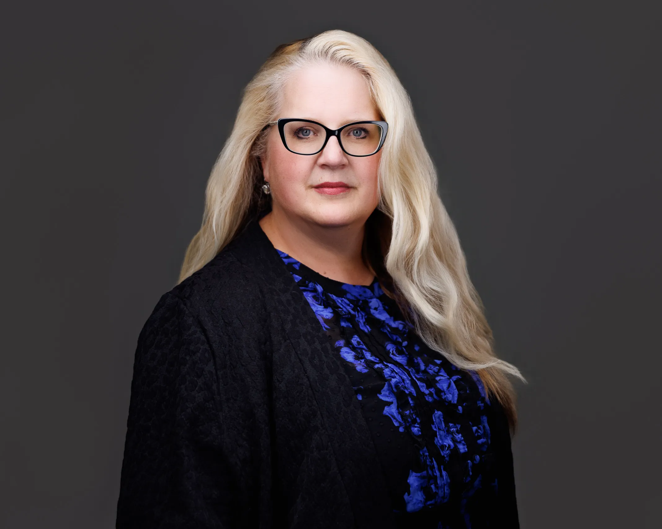 Professional portrait of a blonde woman with glasses, wearing a black jacket and blue patterned blouse.