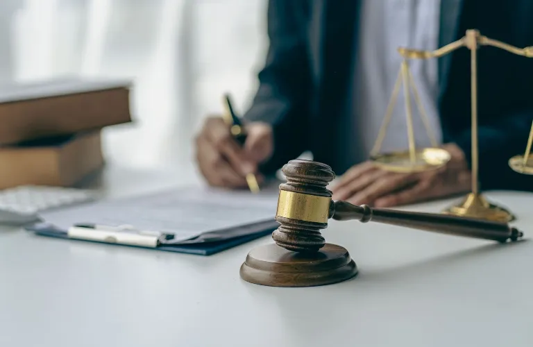 Wooden judge's gavel and brass scales of justice on white desk, person writing in background.