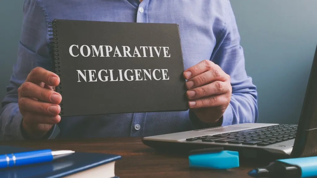 Person in blue shirt holding black notebook with white text "Comparative Negligence" beside laptop and office supplies.