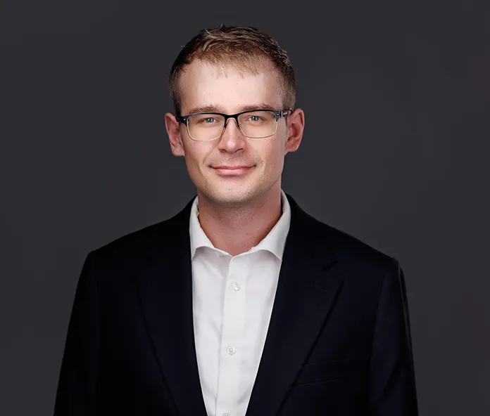 Professional headshot of a young man wearing glasses, a white shirt, and a dark blazer.