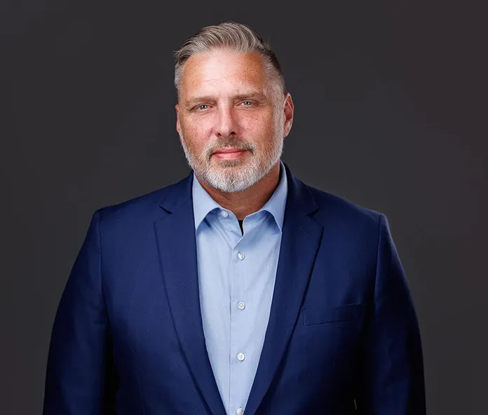 Professional middle-aged man with gray hair and beard, wearing a blue suit and light blue shirt, against a dark gray background.