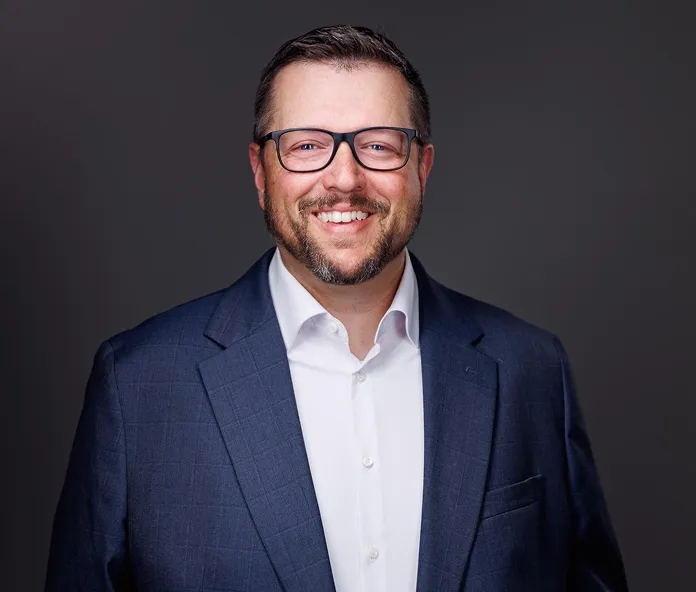 Professional portrait of a smiling man wearing glasses, a navy suit, and a white shirt.