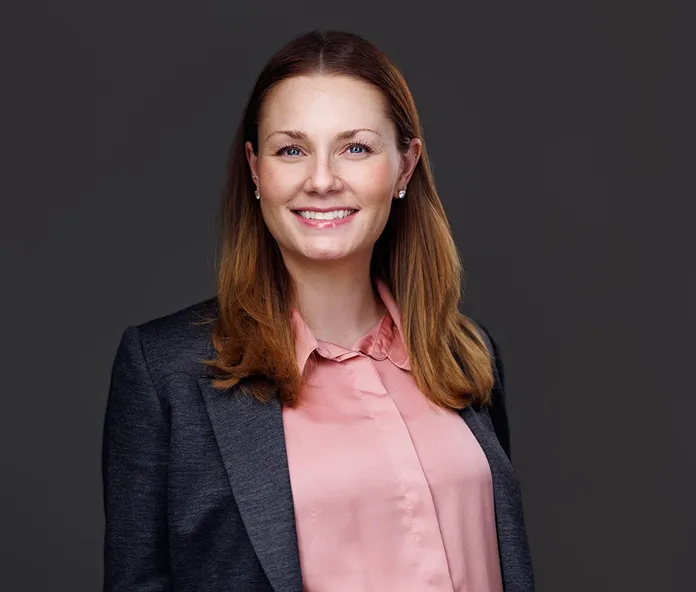 Professional headshot of a smiling woman with light brown hair, wearing a dark blazer and pink blouse.