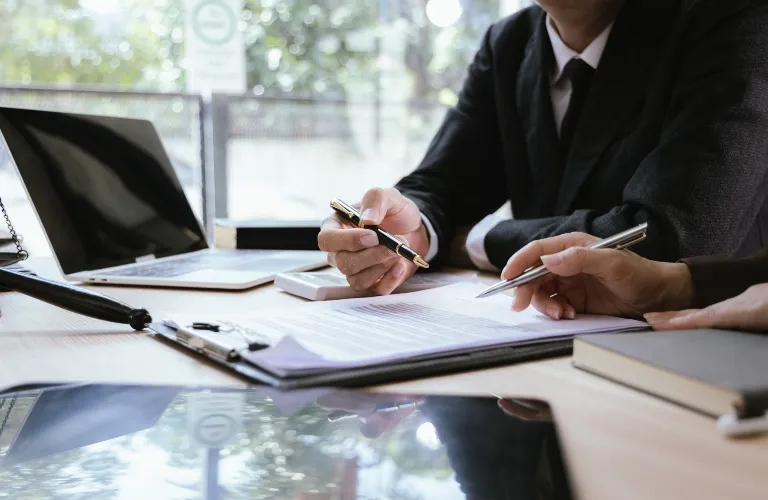 Two professionals in dark suits reviewing documents with pens at a wooden desk.